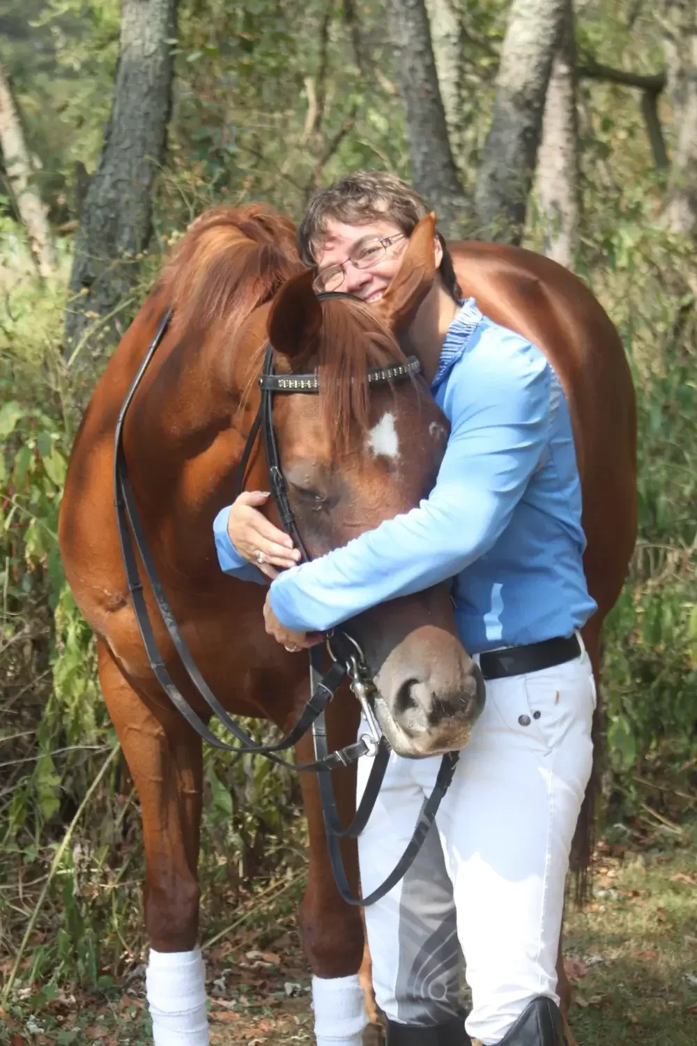 A non-binary person in a blue shirt hugging a brown horse with a white star on its forehead in a wooded area.