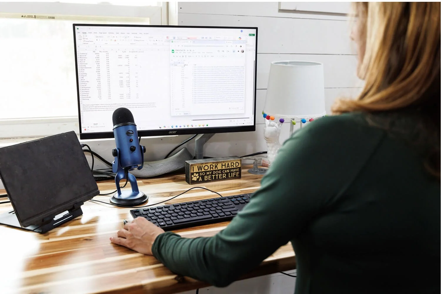 A person working at a desk with a large monitor, a microphone, a keyboard, a tablet, a lamp decorated with colorful pompoms, and a sign that reads 'Work Hard So My Dog Can Have a Better Life'.