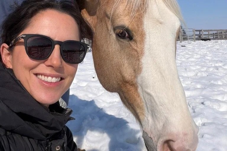 A woman in sunglasses smiling next to a white horse in a snowy field.