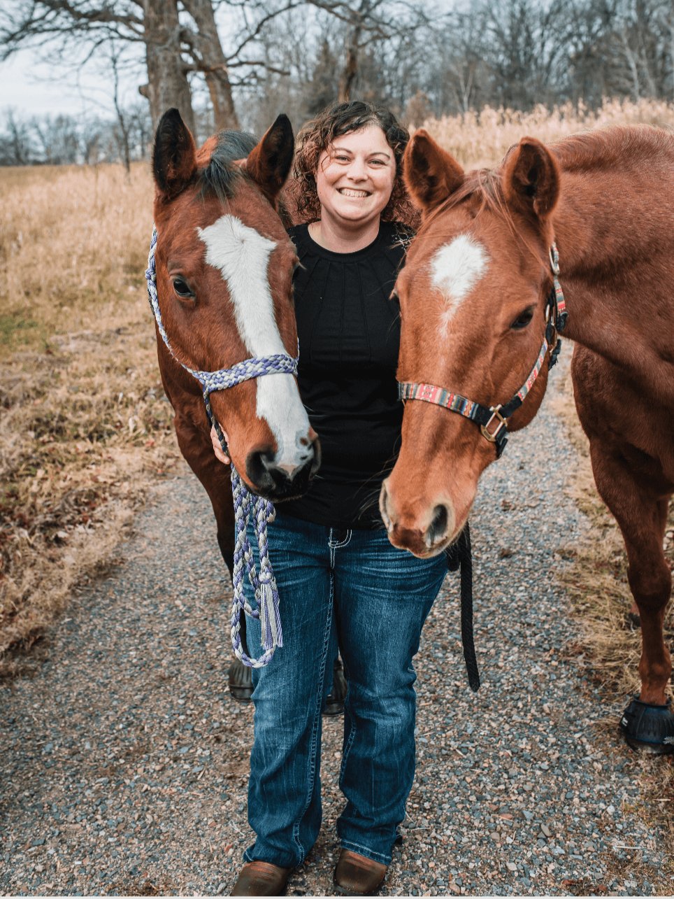 A woman smiling standing outdoors on a gravel path with two brown horses on either side of her.