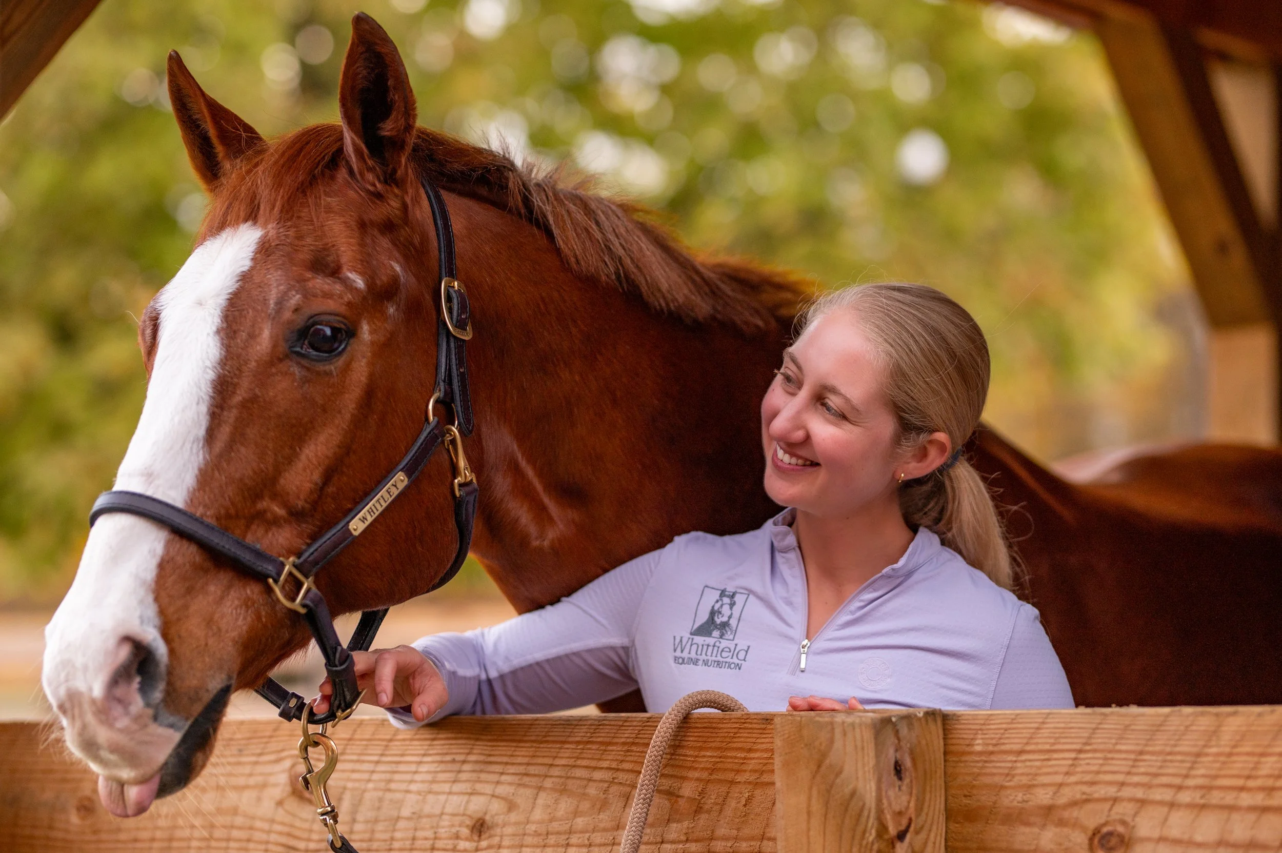 Young woman smiling and interacting with a chestnut horse with a white blaze on its face, standing in a wooden enclosure outdoors, with blurred green trees in the background.
