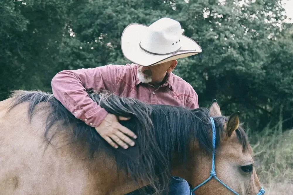 A man wearing a cowboy hat and a red long-sleeve shirt is sitting on a brown horse with a black mane and blue halter, outdoors with trees in the background.