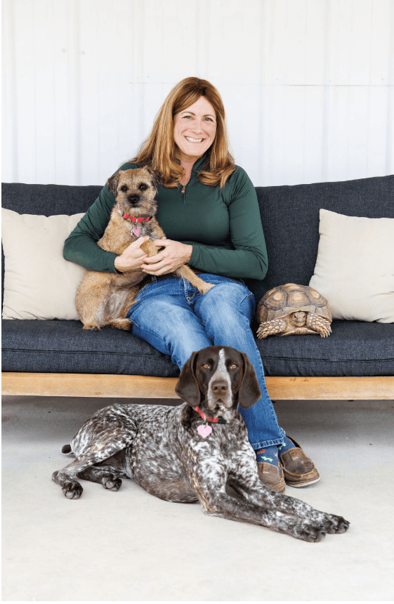A woman sitting on a dark gray couch holding a small dog, with a large dog lying on the floor in front of her, a turtle on the couch beside her, and a white pillow behind her.