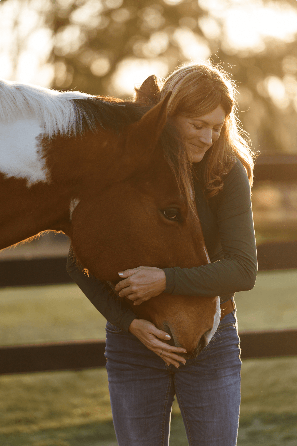 A woman hugging a brown and white horse outdoors in warm sunlight, with trees blurred in the background.