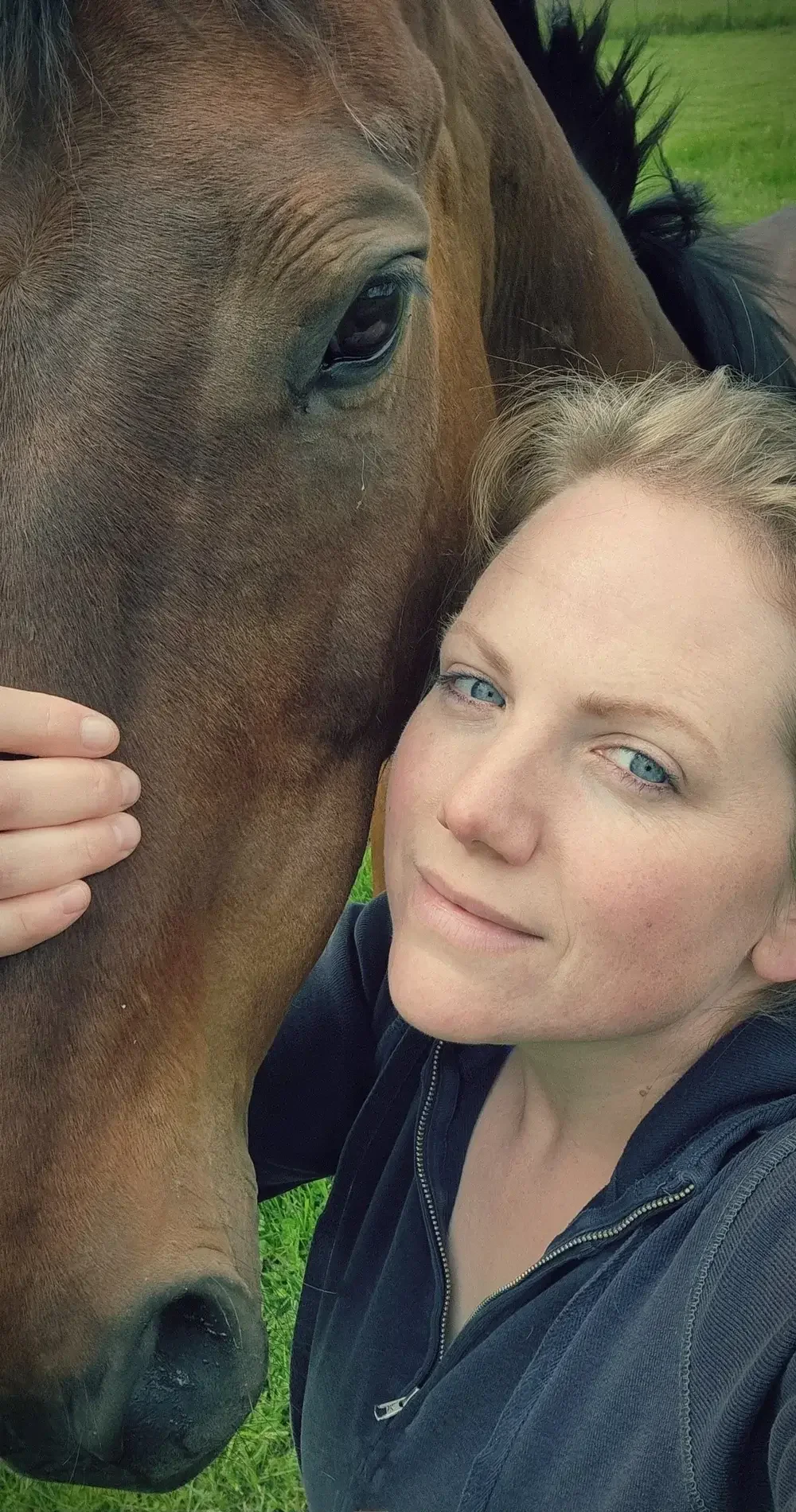 A woman with blonde hair and blue eyes taking a close-up selfie with a brown horse, both appearing calm and friendly outdoors on a grassy field.