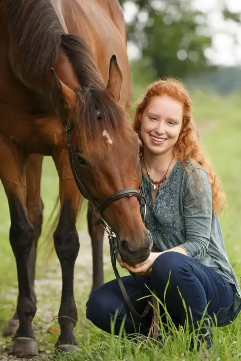 A woman with red hair kneeling beside a brown horse outdoors in a grassy area, smiling and gently holding the horse's muzzle.