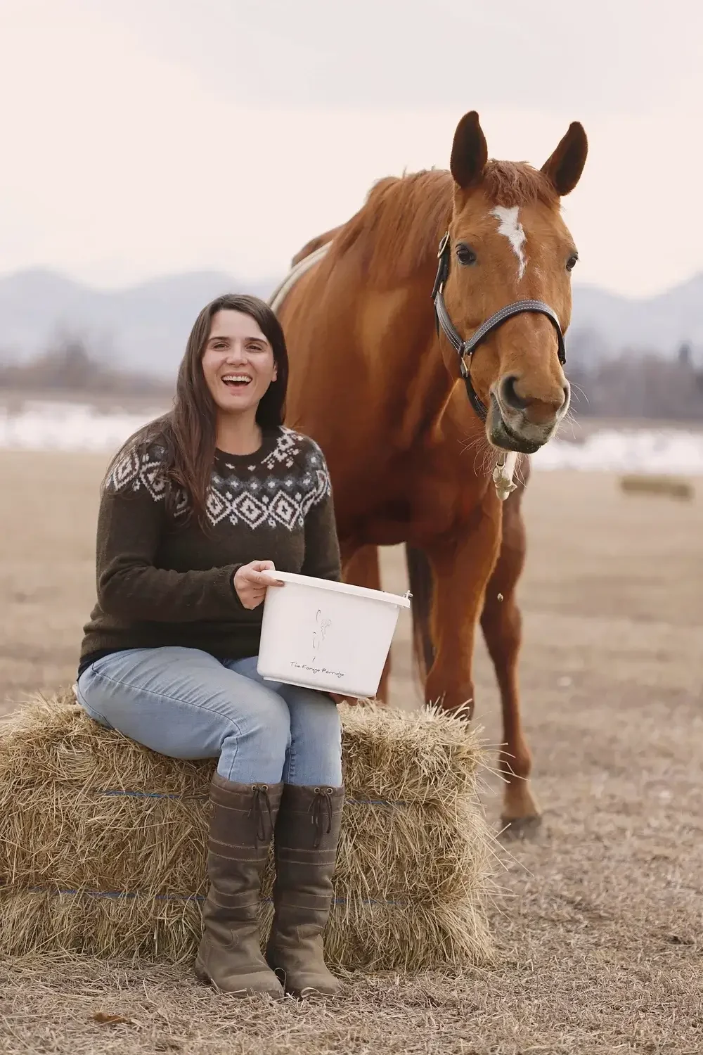 A woman sitting on a hay bale holding a white box, smiling, with a brown horse behind her in an outdoor farm setting.