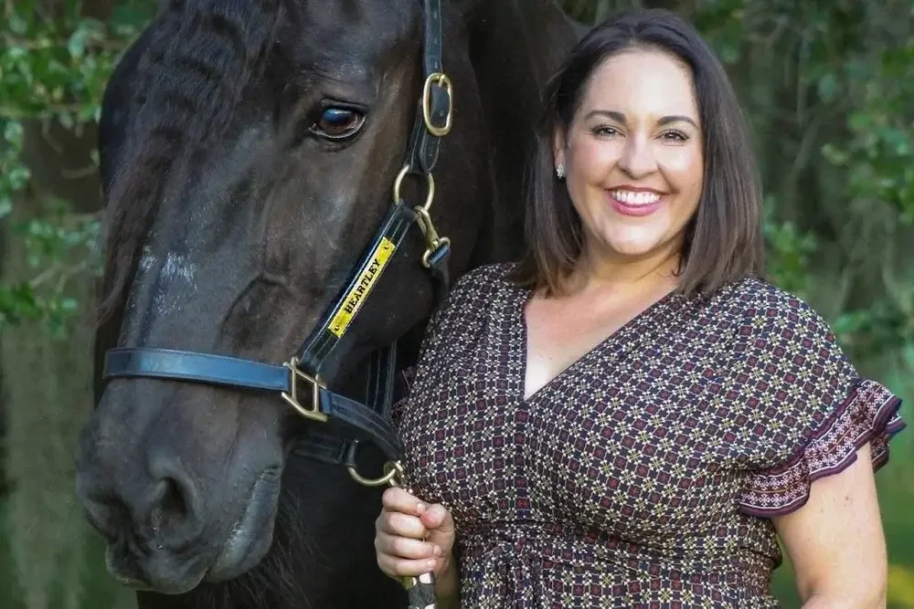 A woman standing next to a black horse outdoors, holding its lead. The woman is smiling and has shoulder-length dark hair, wearing a patterned dress. The horse has a halter with blue and gold details, and the background features green foliage.