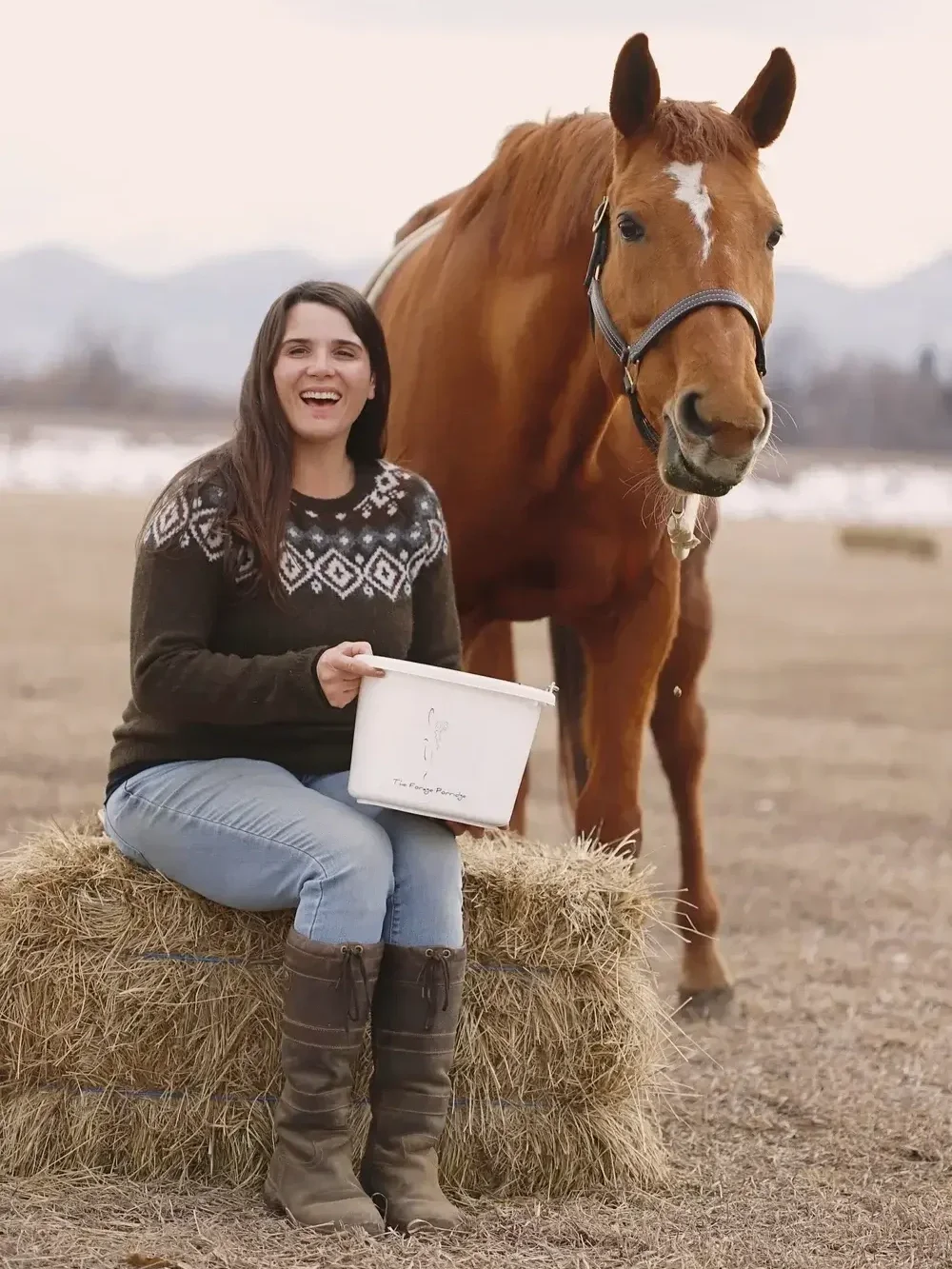 A young woman sitting on hay bales outdoors next to a brown horse with a white star marking on its forehead. She is smiling, wearing a black patterned sweater, blue jeans, and brown boots, holding a white box with a horse illustration and text.