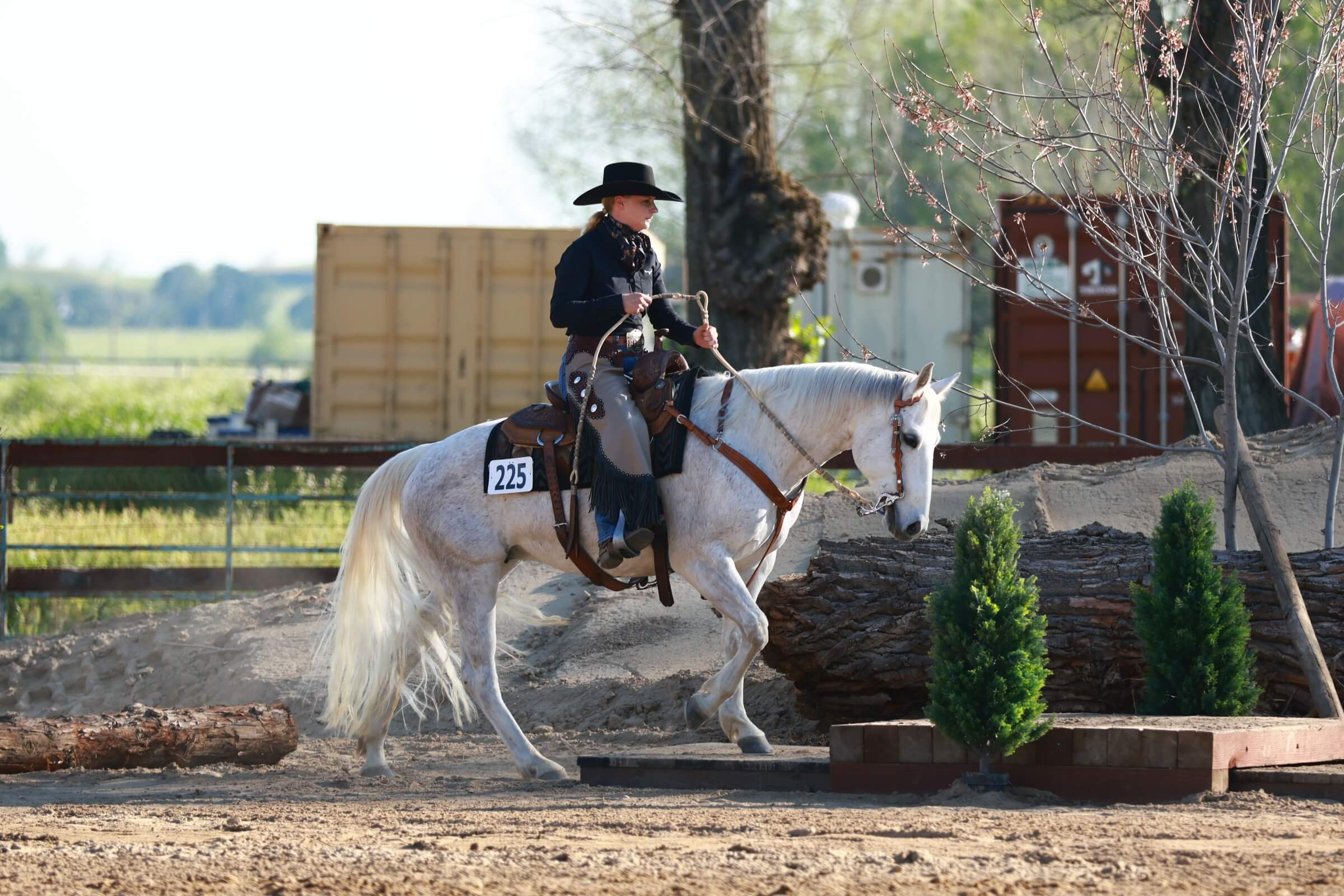 A woman dressed in western attire riding a white horse in an outdoor, natural setting with trees and a riding obstacle.