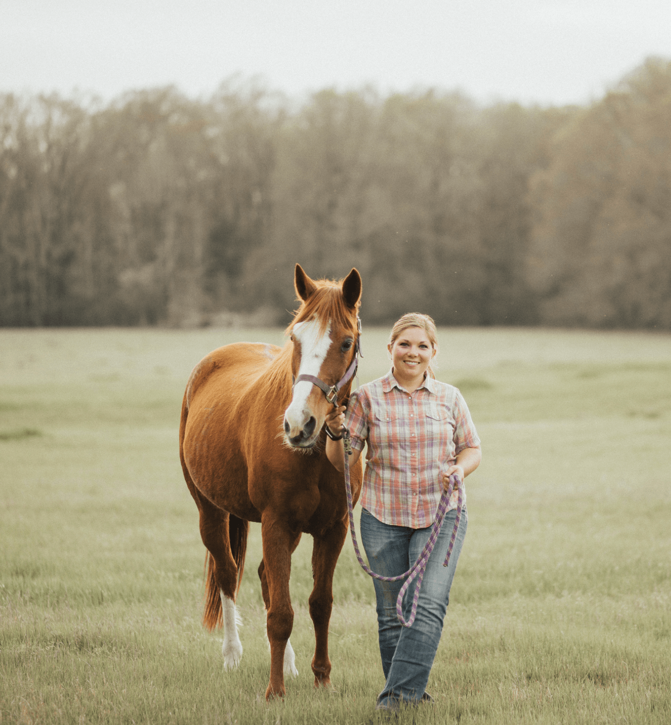 A happy young woman walking on a lush green field, holding a purple lead rope attached to a brown and white horse, with a background of trees.