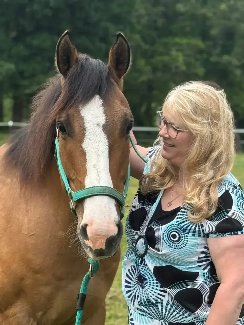 A woman with blonde hair, glasses, and a patterned shirt standing beside a brown horse with a white stripe on its face, outdoors with green trees in the background.