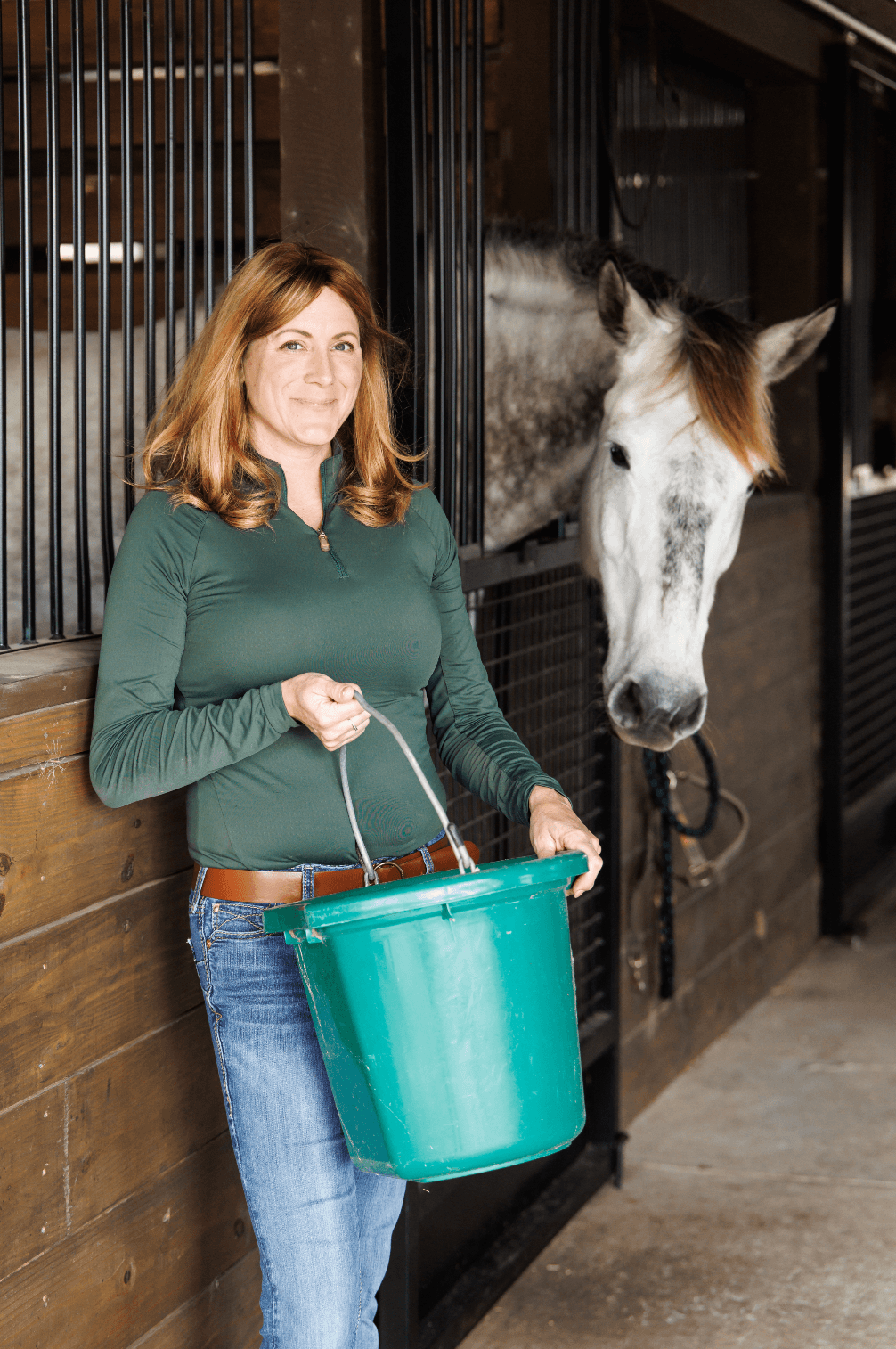 A woman standing in front of a horse stall inside a barn, holding a green buckets, while a white and gray horse leans its head out of the stall.