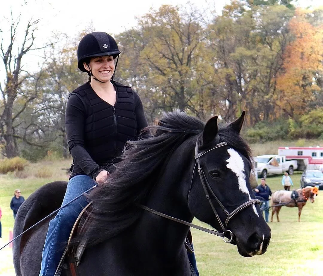 A woman riding a black horse with a white blaze on its face in an outdoor setting with autumn trees and people in the background.