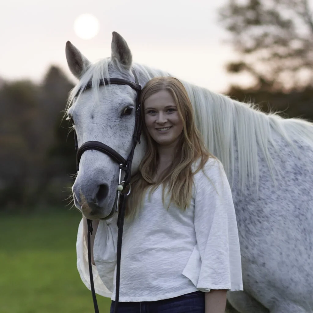 A woman with long red hair smiling next to a white horse outdoors at sunset.