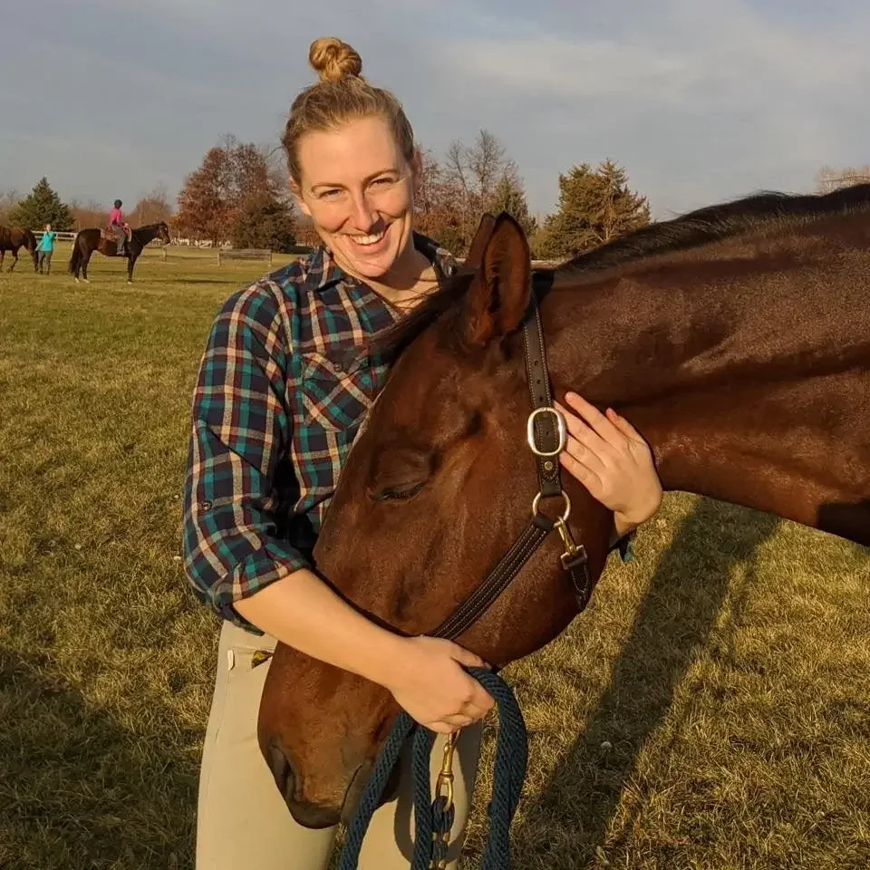 A woman in a plaid shirt is hugging a brown horse outdoors on a grassy field, with a few people riding horses in the background.