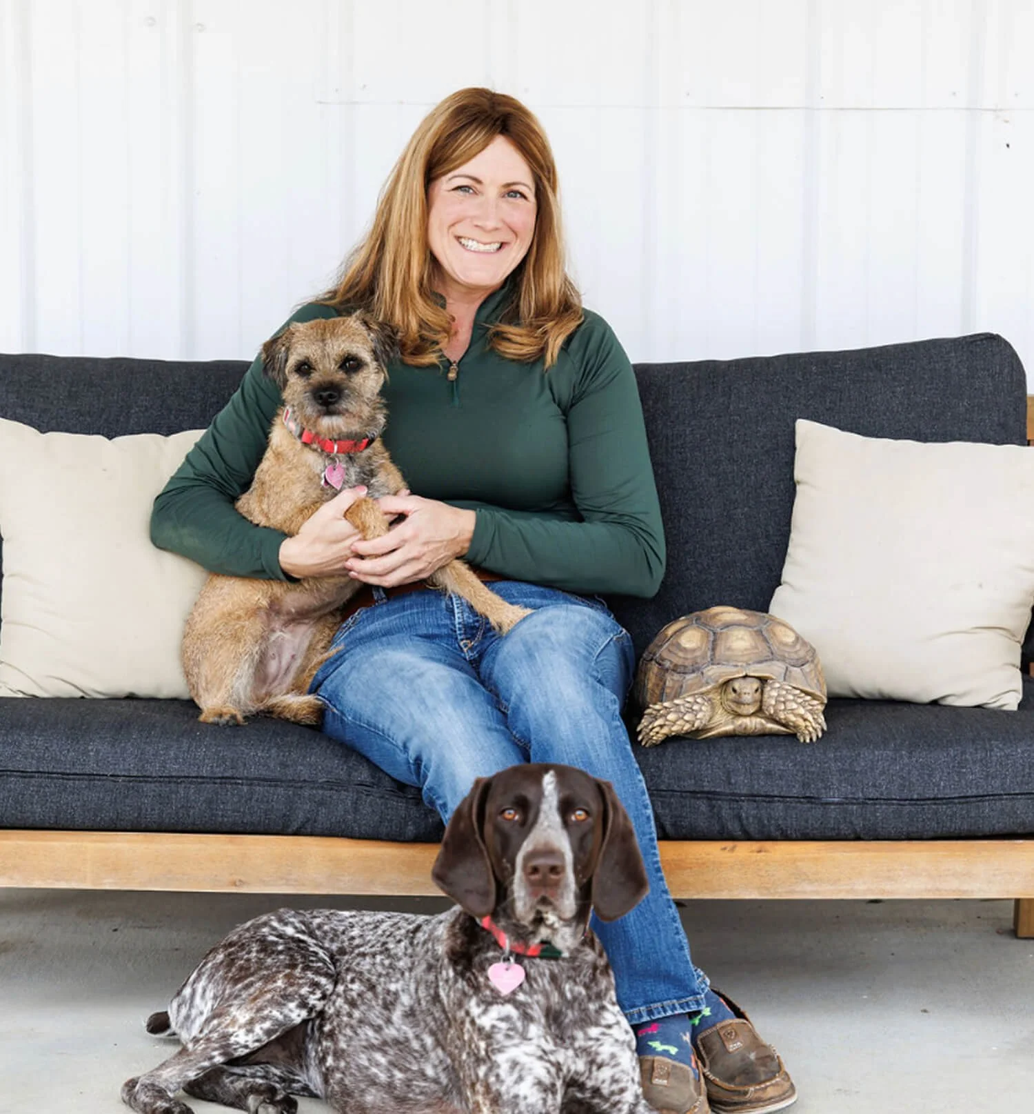 Woman sitting on a black sofa holding a small dog with brown and black fur. A large decorative turtle statue is beside the woman. A brown and white dog is sitting on the floor in front of her, and a brown and white dog with a pink heart tag is lying on the floor nearby.