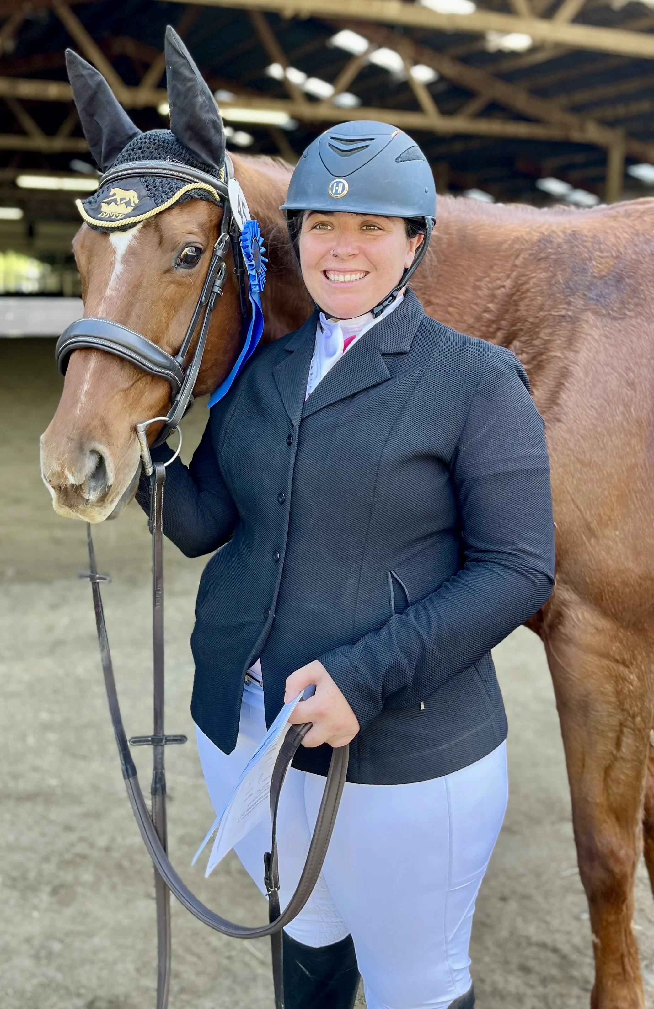 A smiling woman dressed in equestrian attire, including a helmet, jacket, and white riding pants, standing next to a brown horse with a ribbon on its bridle in a riding arena.
