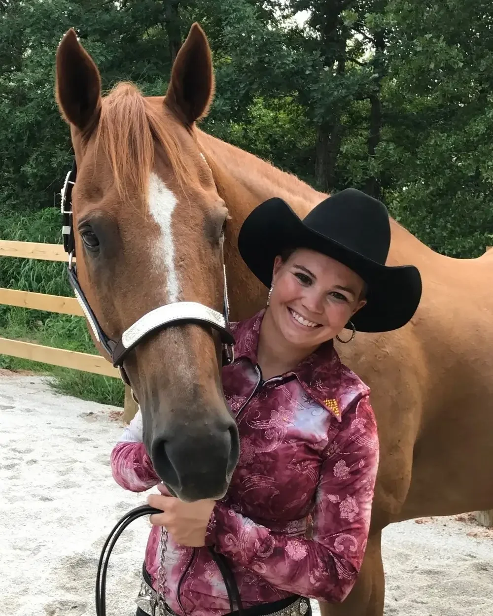 A smiling woman in a black cowboy hat and floral western shirt standing with a brown horse, outdoors with trees and a fence in the background.