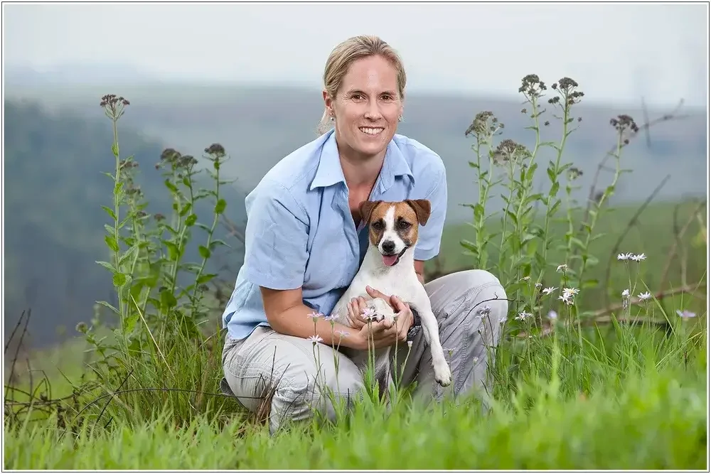 A woman in a light blue shirt crouching in a grassy field, holding a small brown and white dog. The background shows a blurred landscape with green hills and a cloudy sky.