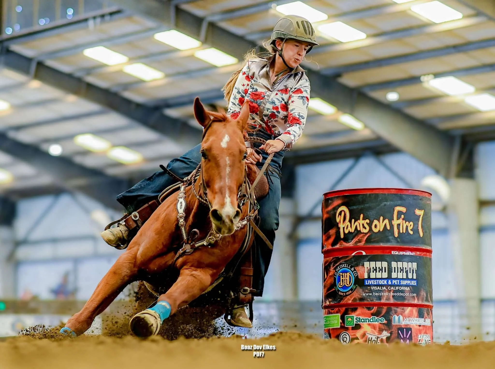A woman riding a horse at a rodeo arena wearing a helmet and floral shirt, jumping over a barrel with feed depot branding, with a focus on her determined expression and the horse's movement.