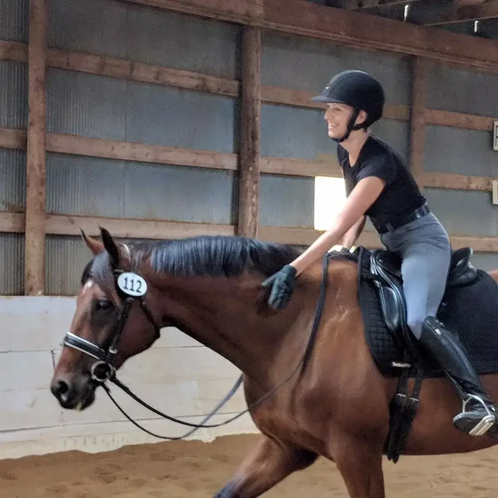 A woman riding a brown horse inside an indoor riding arena, wearing a black helmet, black shirt, gray riding pants, and gloves.