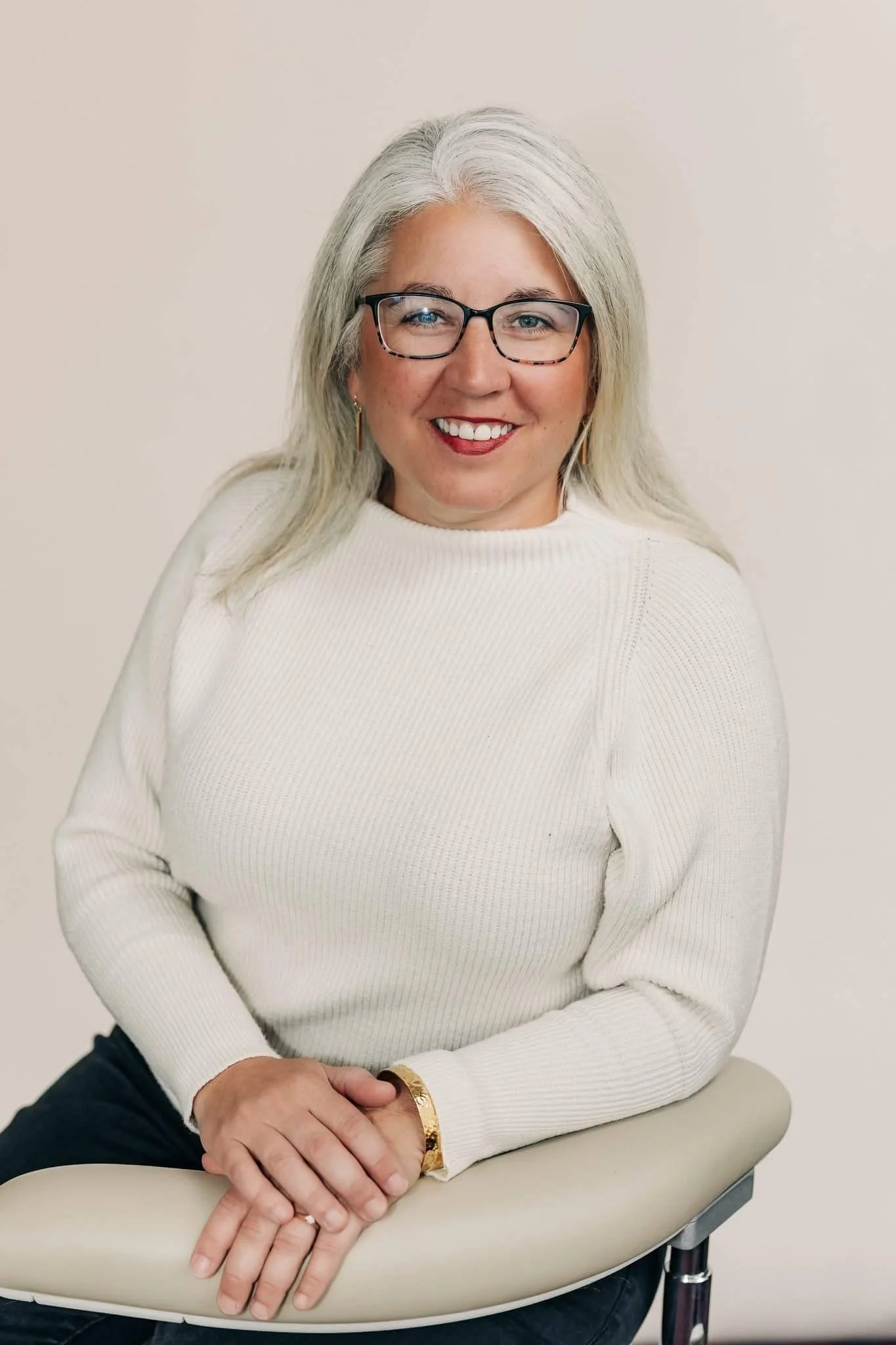 Woman with gray hair, glasses, and hoop earrings sitting on a chair, smiling, wearing a white sweater and gold bracelet.