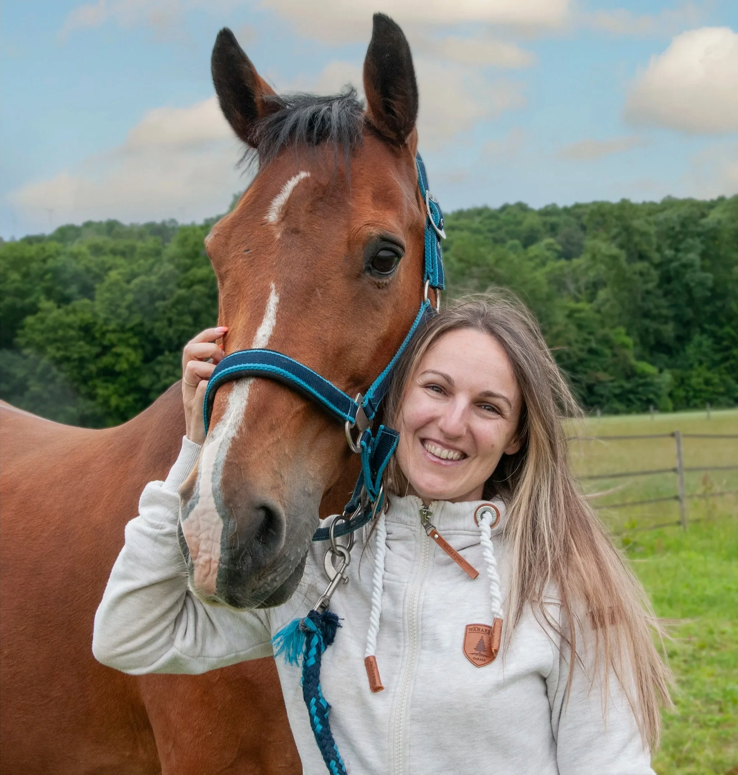 A woman with blonde hair smiling while holding a brown horse's head. The horse has a white star-shaped mark on its forehead and is wearing a blue halter. The background features green trees and a cloudy sky.