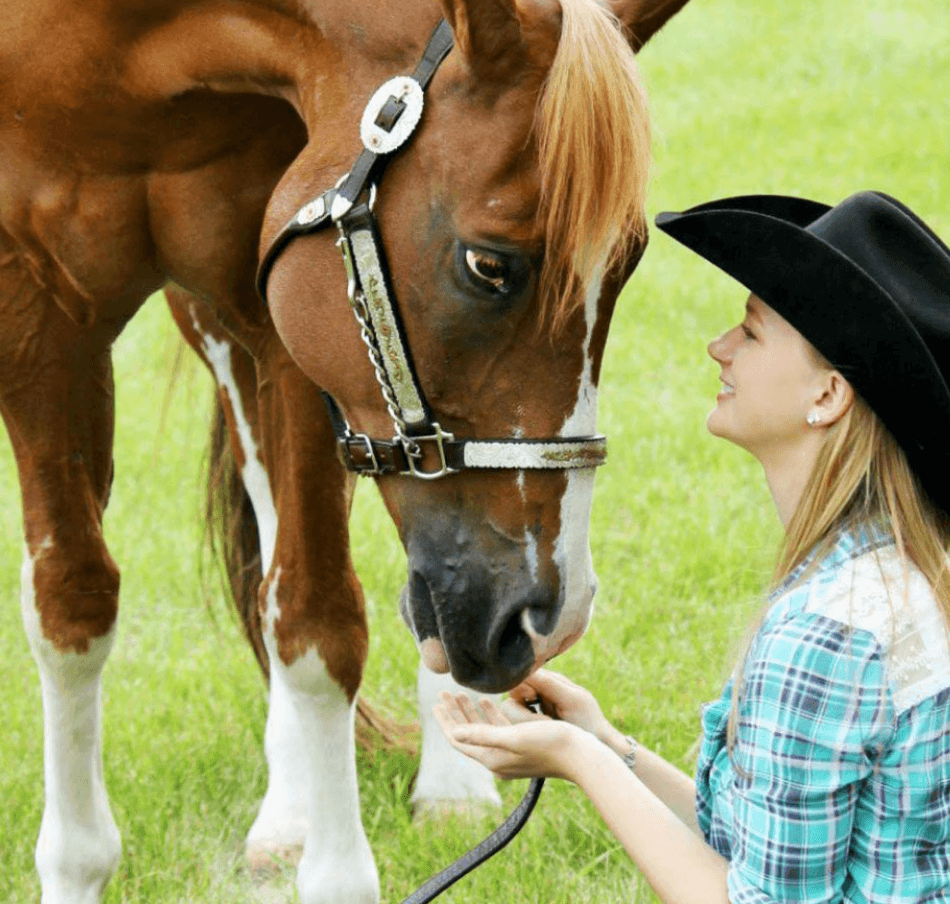 A woman wearing a black cowboy hat and plaid shirt gently touching a brown and white horse's face in a grassy field.