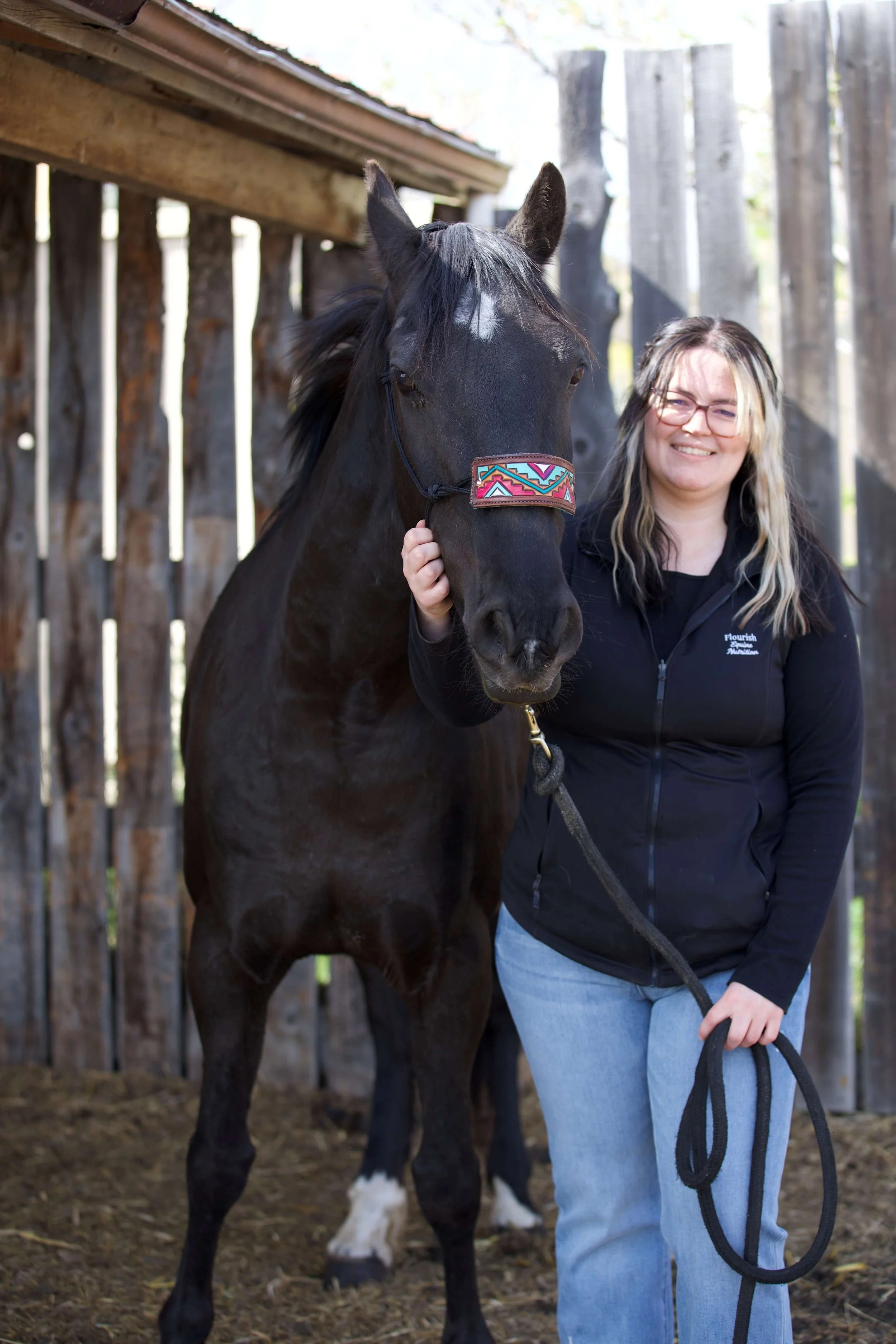 A woman wearing glasses and a black jacket standing next to a black horse with a patterned halter, in an outdoor wooden fenced area with sunlight filtering through trees.