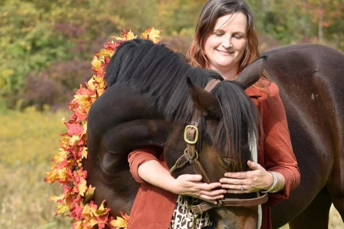 A woman hugging a brown horse wearing a colorful autumn leaf garland around its neck, outdoors with fall foliage in the background.