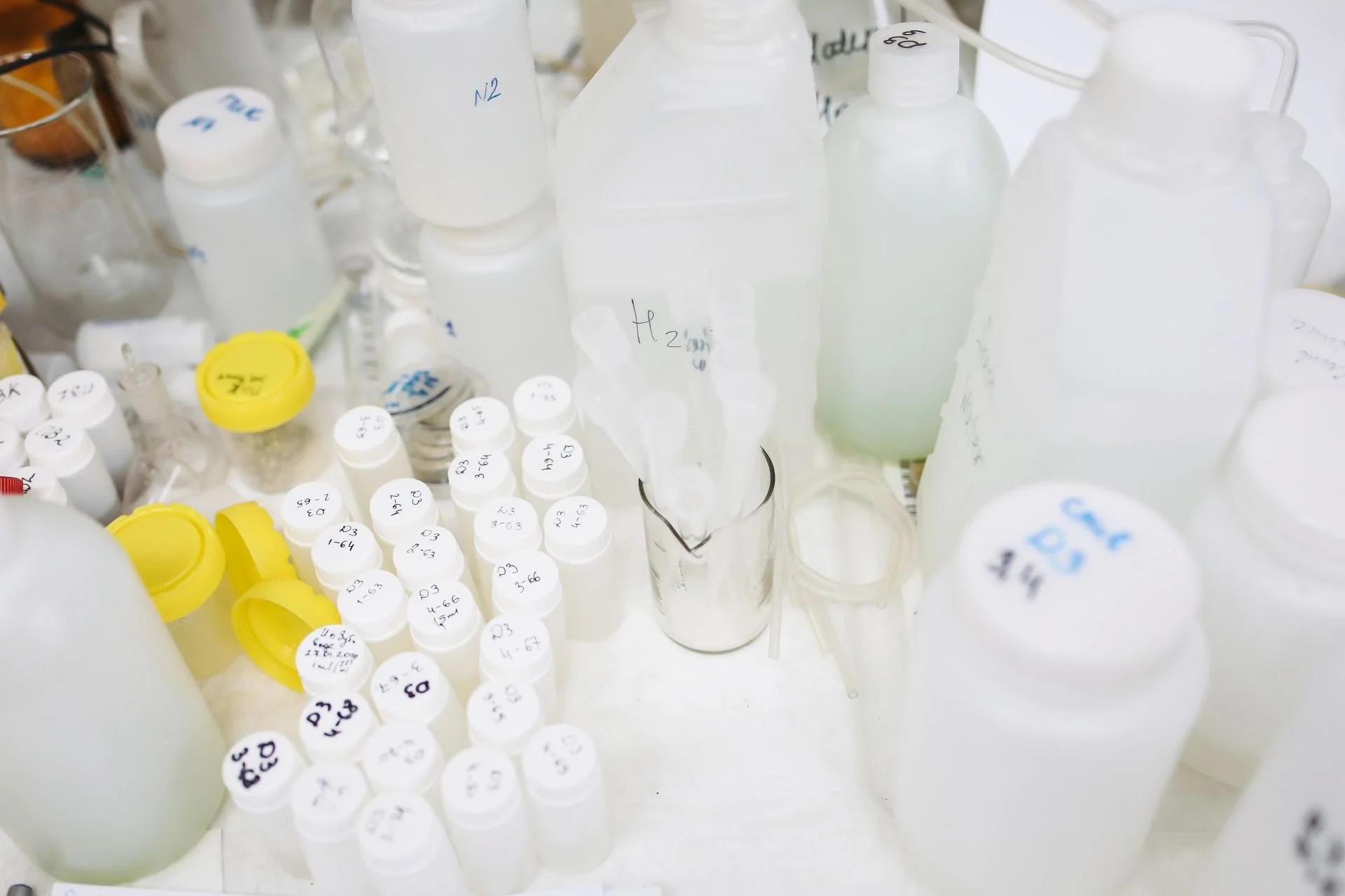 White laboratory bottles, labeled sample containers, pipettes, and testing equipment arranged on a lab bench for chemical analysis.