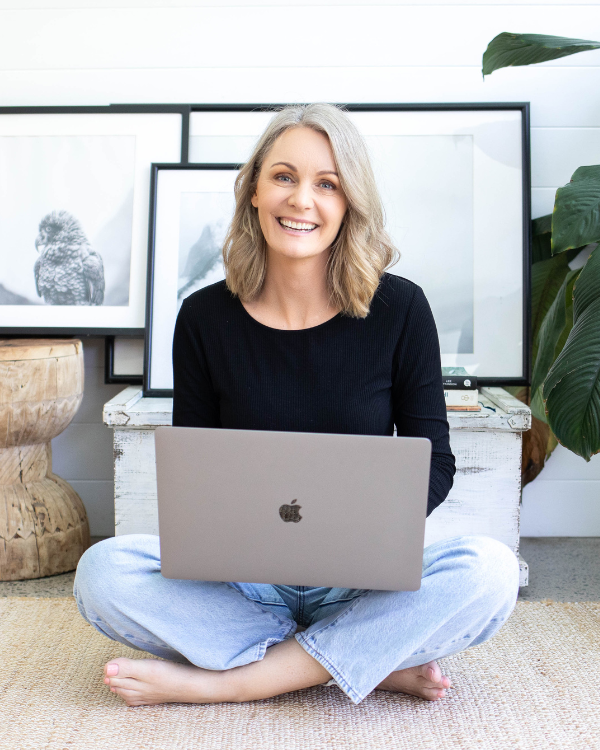 Kate, Squarespace website designer and owner of Siya Studio sitting cross-legged on a beige carpet with a silver laptop on her lap, smiling in a bright living room with framed black-and-white bird photographs and a large green plant behind her.
