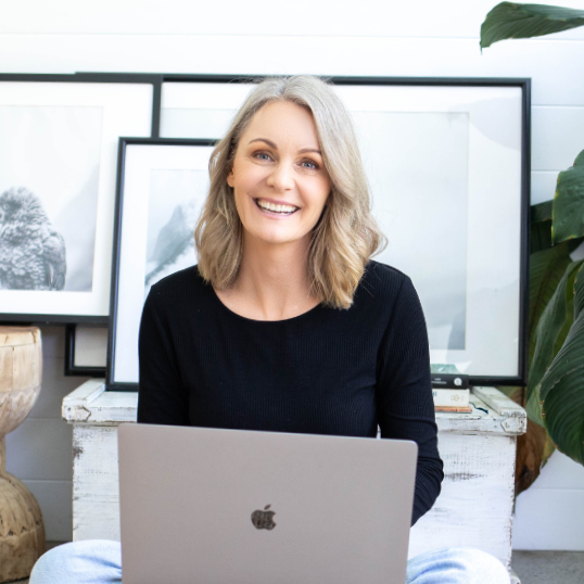 A smiling woman sitting in front of a laptop with framed art and houseplants in the background.