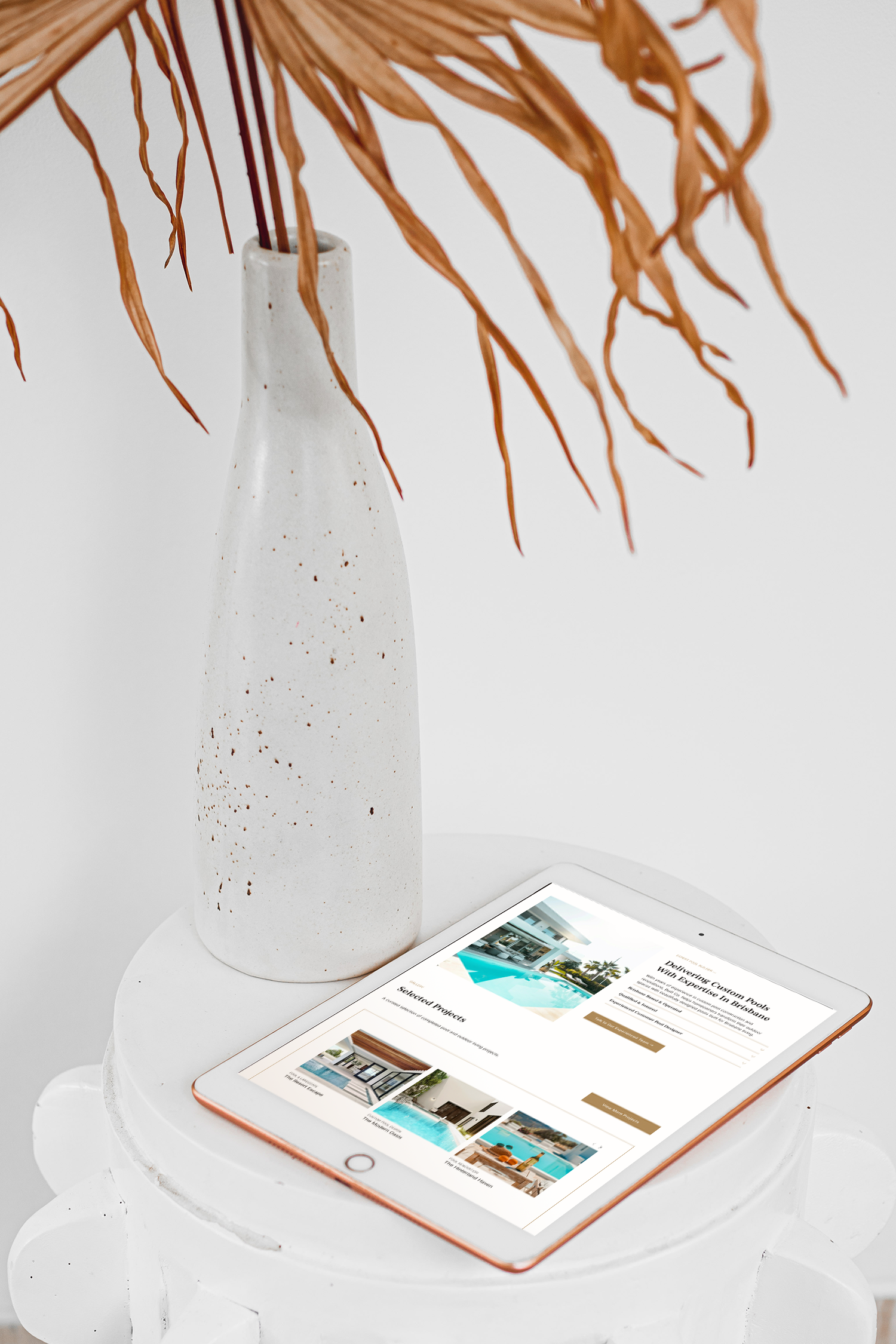 A white ceramic vase with speckled pattern holding dried orange-brown palm leaves, placed on a white round table next to a white tablet displaying a modern and sleek portfolio for real estate website.