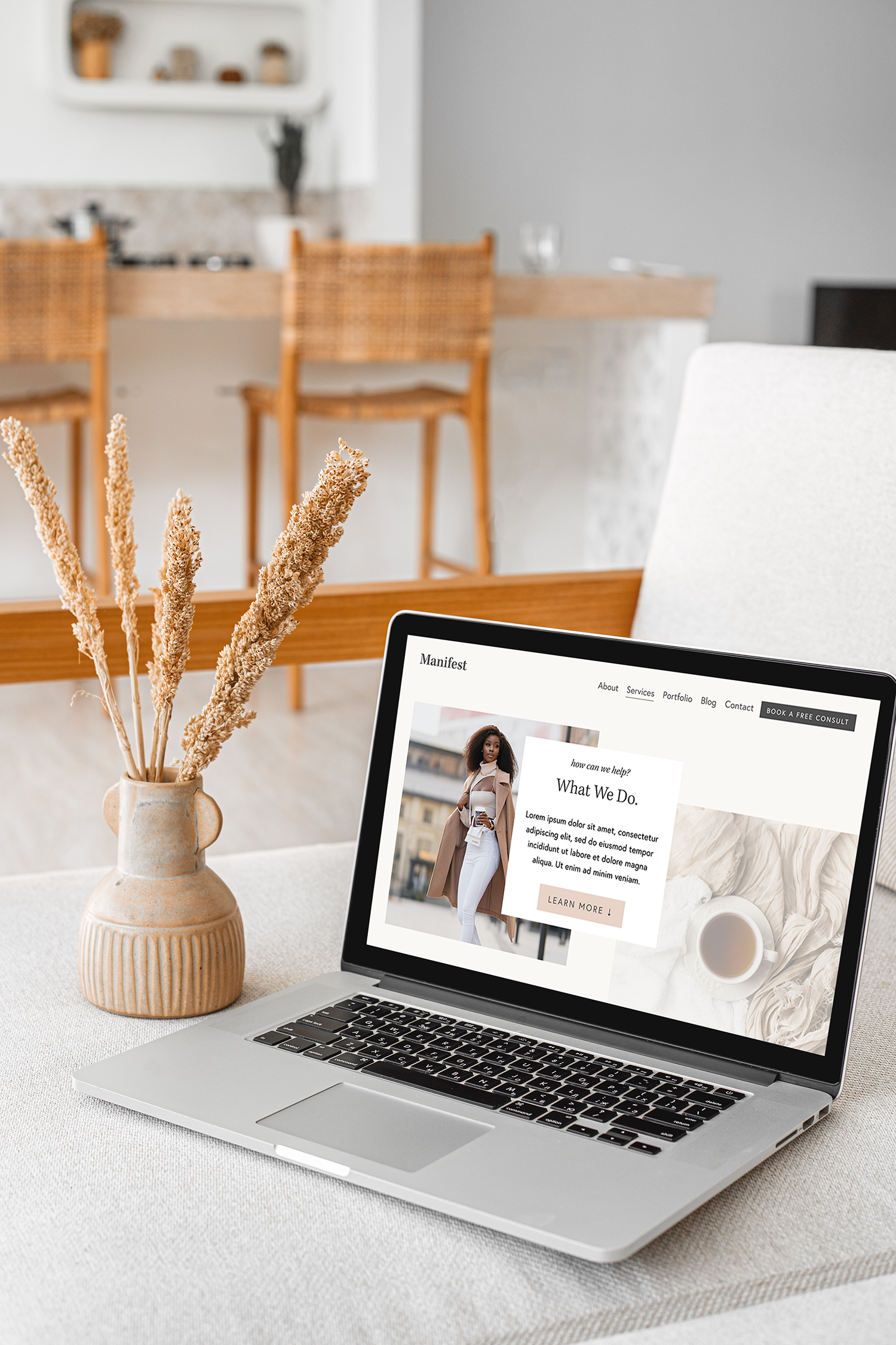 Open laptop displaying a website with a woman on it, placed on a table with a beige tablecloth. Next to the laptop is a ceramic vase with dried plants. In the background, there is a wooden dining table with chairs and a partial view of a kitchen area.