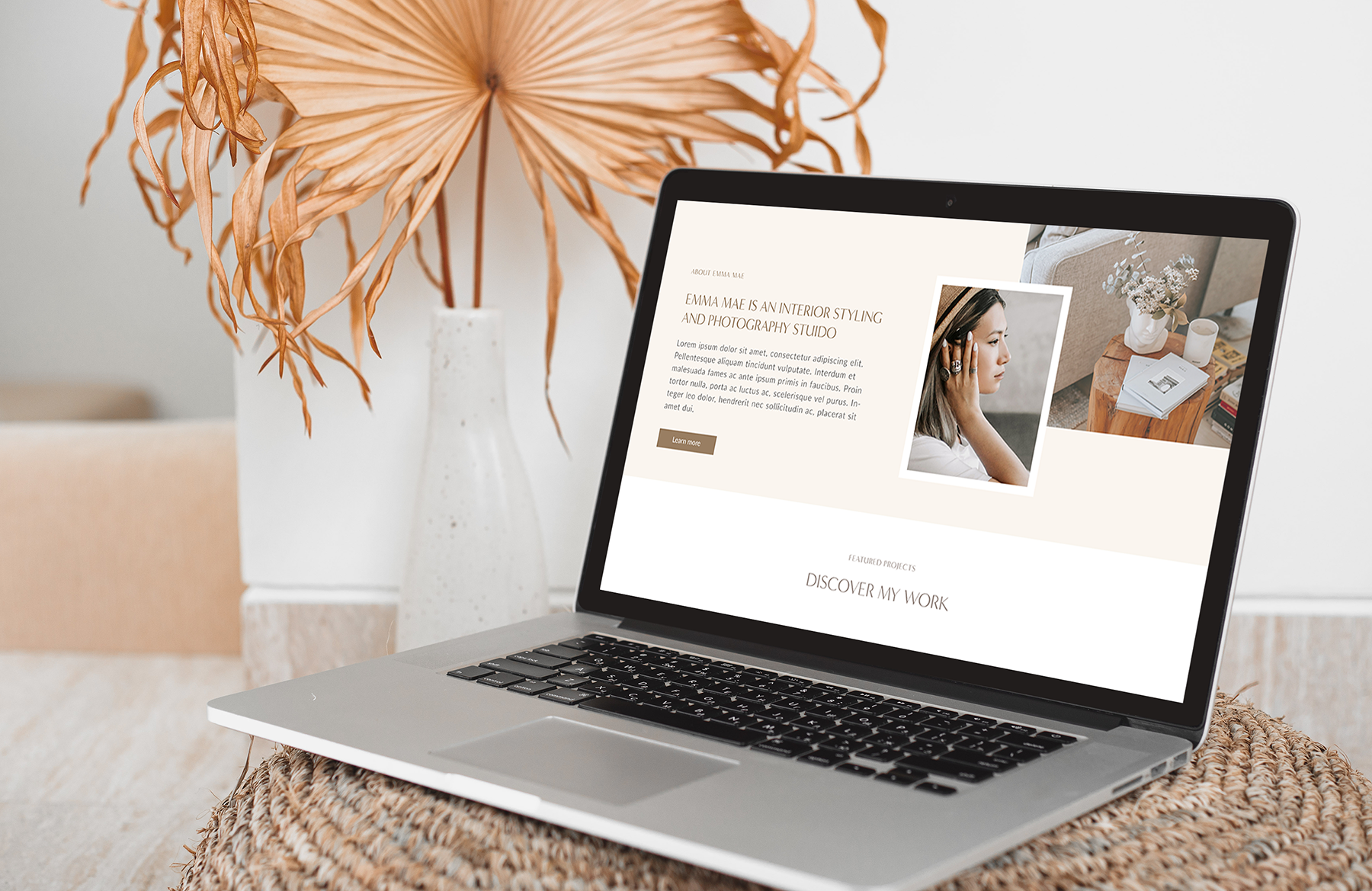 A laptop displaying a clean and modern website design for interior styling and photography, design by Siya Studio, placed on a woven surface with a vase of dried leaves in the background arranged in a white vase.