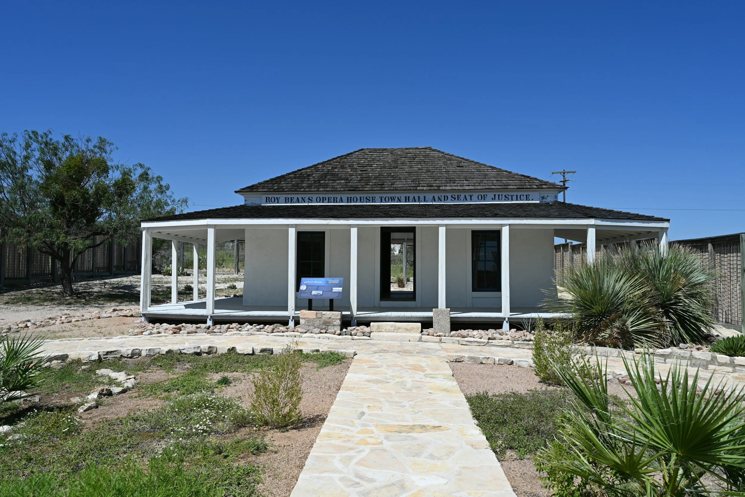 White building with a porch and shingle roof, sign reads Roy Bean's Opera House Town Hall and Seat of Justice, surrounded by desert plants and a stone walkway, under a clear blue sky.