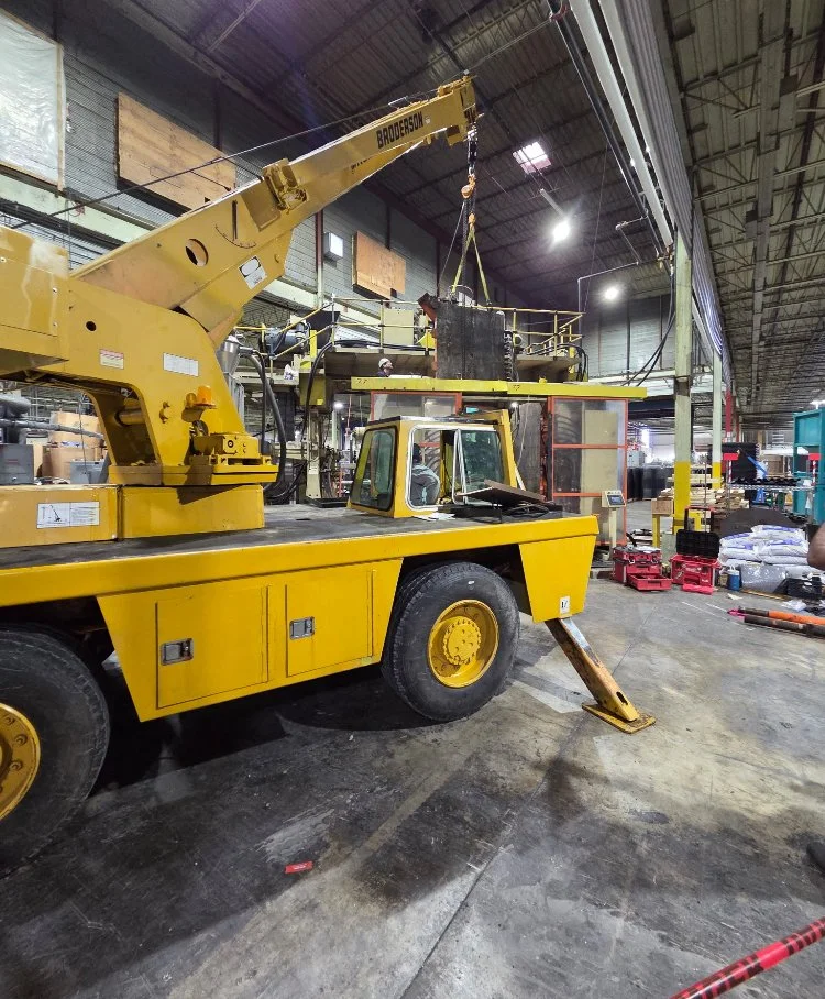 Yellow crane inside an industrial warehouse, with a long extendable arm and a black lifting hook, surrounded by pallets and equipment.