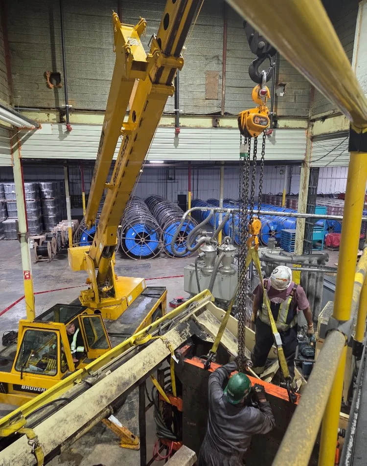 Workers operate a yellow crane to lift a large electrical transformer inside an industrial warehouse, with spools of wire or cable stacked against the wall in the background.