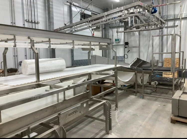Industrial food processing machinery in a clean factory setting, including conveyor belts, rollers, and metal frames.
