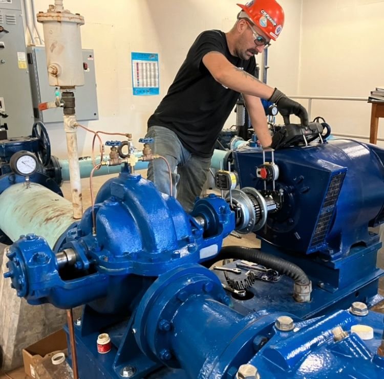 A technician wearing a red safety helmet, glasses, and gloves working on industrial machinery, including a large blue pump or motor, in a mechanical room.