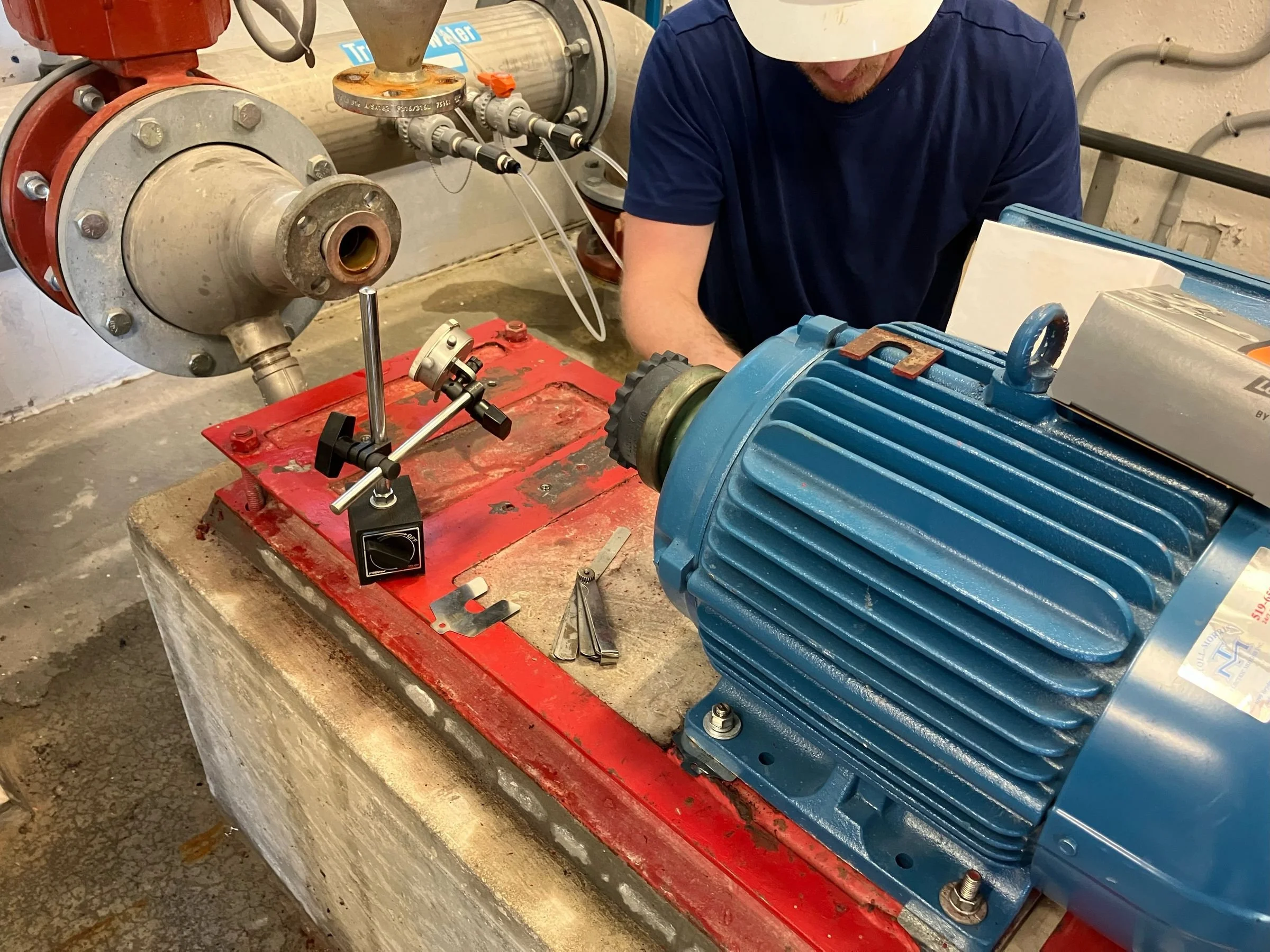 A worker in a white hard hat and dark blue shirt is working with industrial machinery, including a large blue electric motor connected to a red pipe with bolts and a metal flange. The environment appears to be a processing or manufacturing plant with