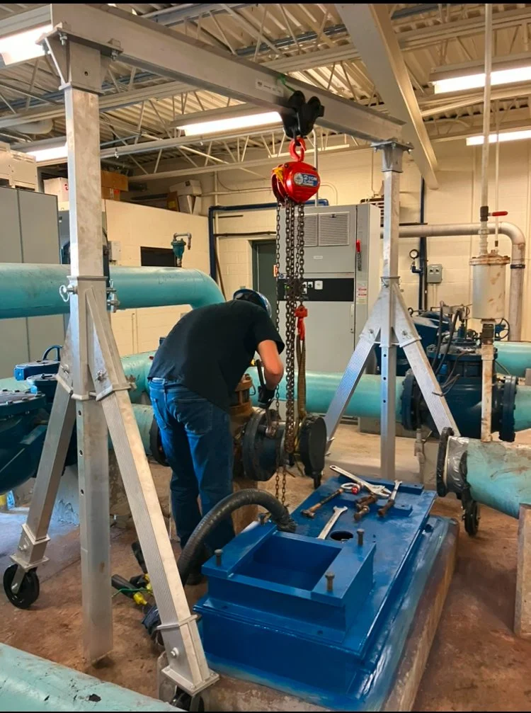 A person working on industrial equipment in a factory, using tools around large blue pipes and machinery, with a chain hoist and overhead crane in the workspace.