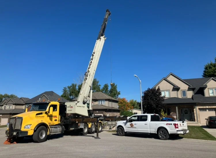 A crane truck lifting a utility pole in a residential neighborhood with houses and trees under a clear blue sky.