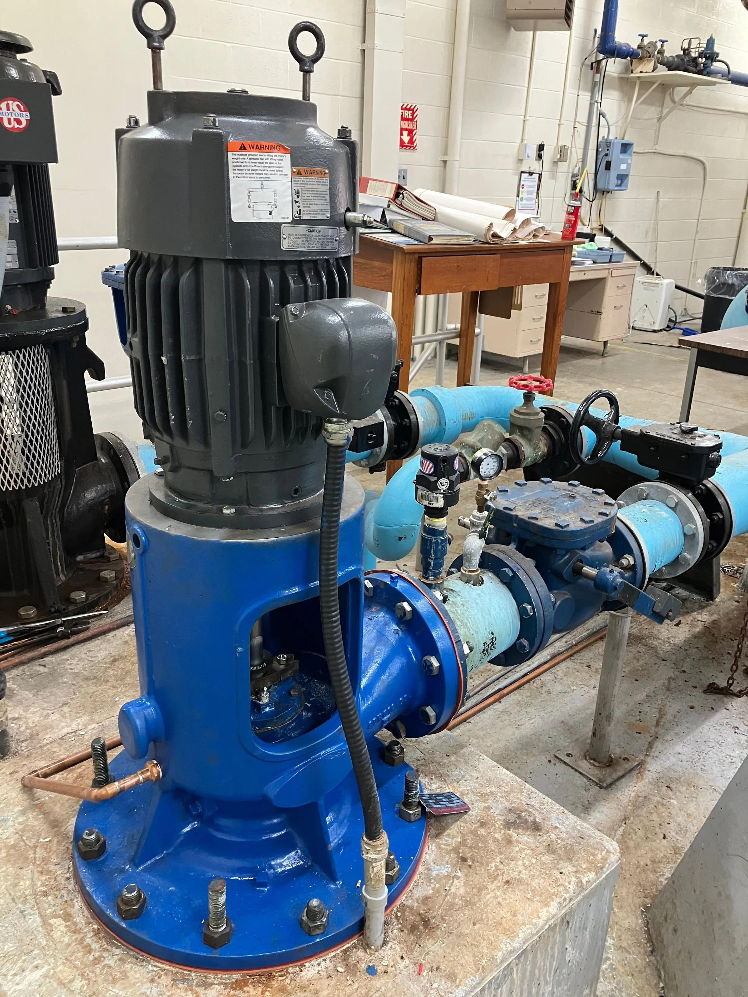 A large industrial water pump with blue casing and interconnected piping, installed on a concrete platform in a utility room.