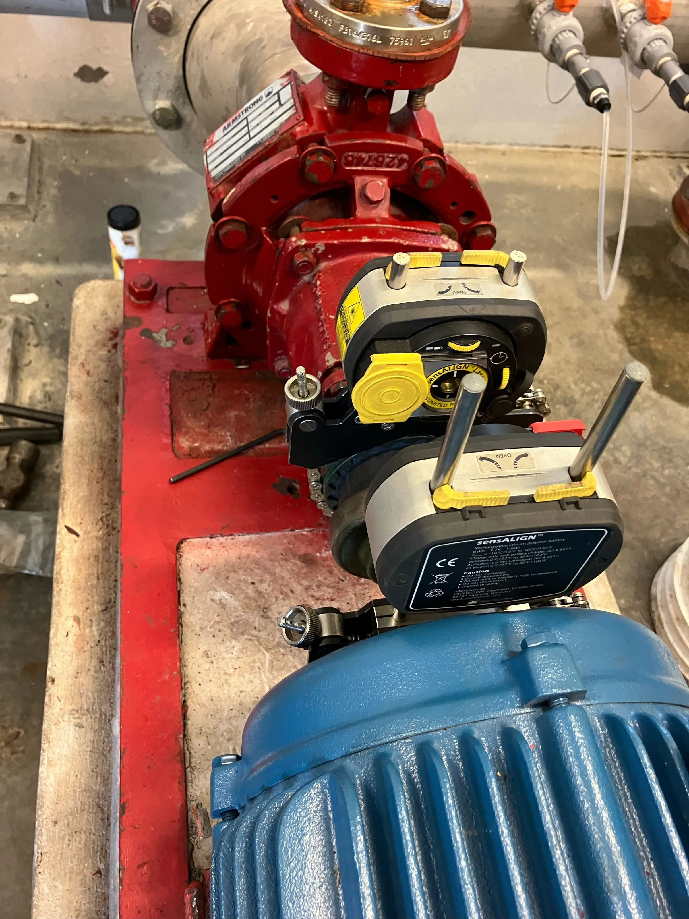 Close-up of a mechanical setup with a blue pipe, a red pump, a black and yellow device, and some wires, on a red work surface in a workshop or industrial setting.