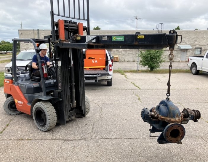 A worker operating a forklift that is lifting a large, rusty pipe fitting in an outdoor parking lot.