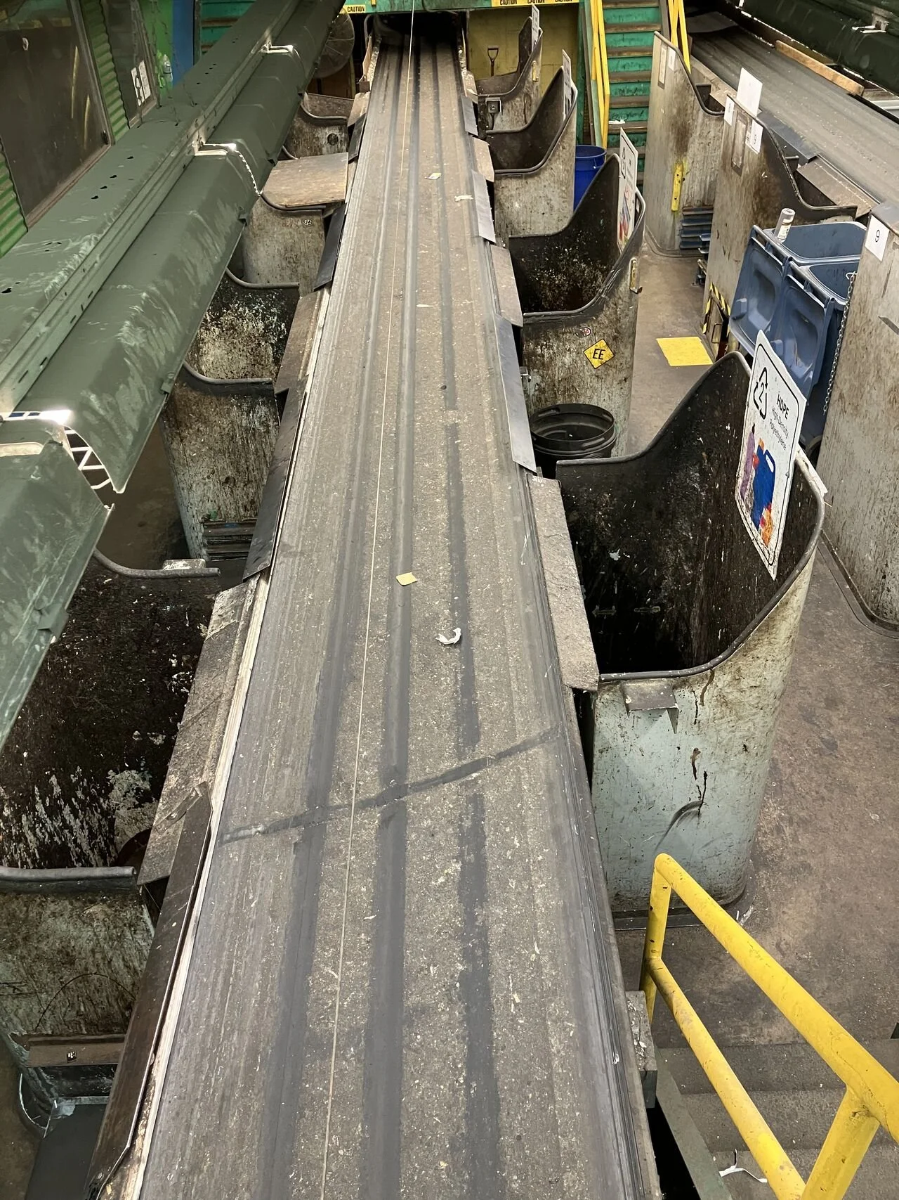 Empty, dirty conveyor belt in an industrial setting with food service signage, surrounded by rusted metal bins and yellow safety barriers.