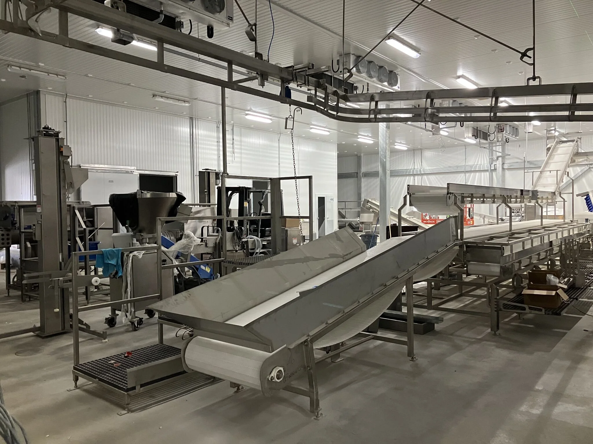 View of an industrial facility or processing plant with metal equipment, conveyor belts, and overhead piping, under bright fluorescent lighting.